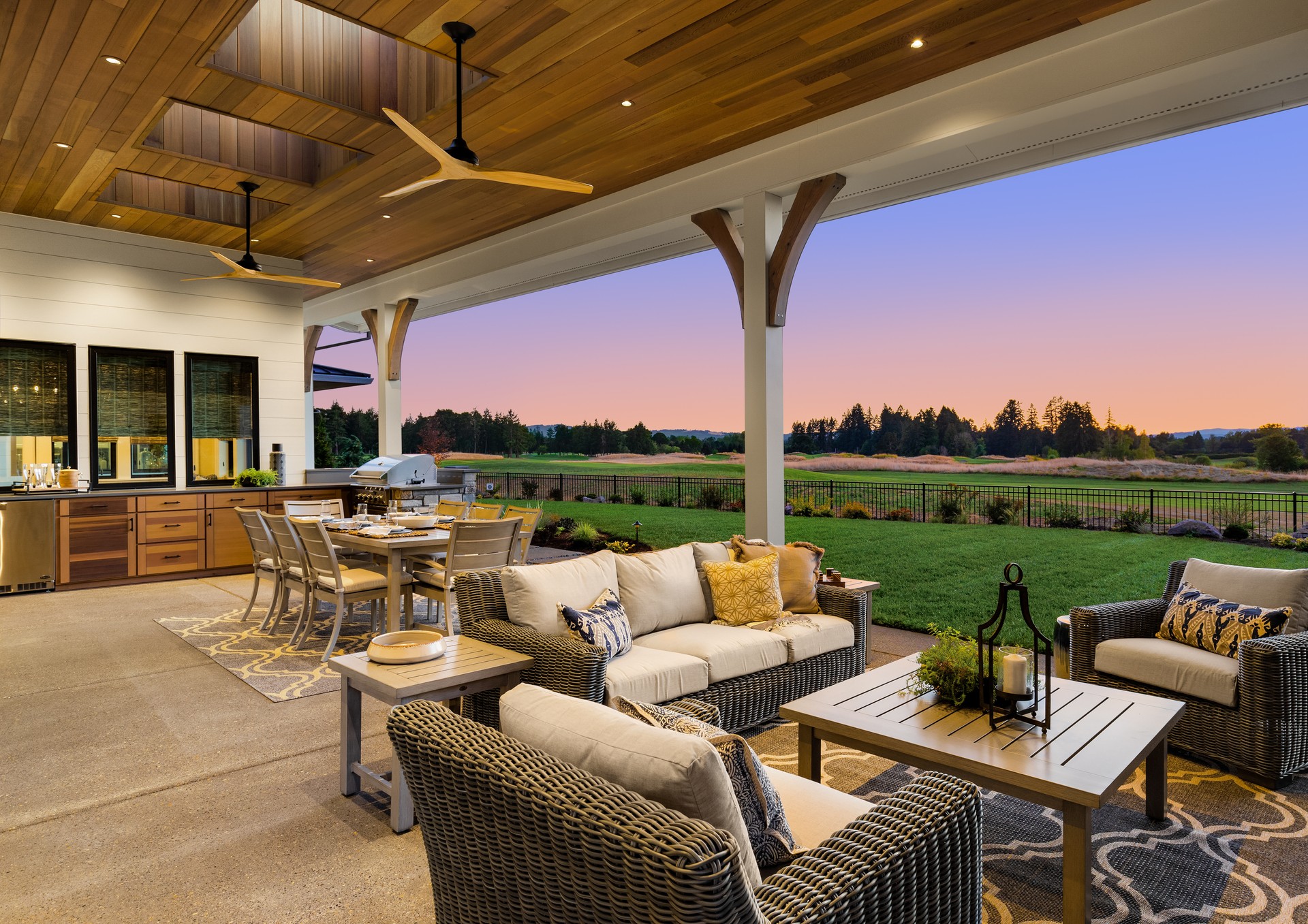 Luxury home exterior at sunset: Outdoor covered patio with kitchen, barbecue, dining table, and seating area, overlooking grass field and trees.